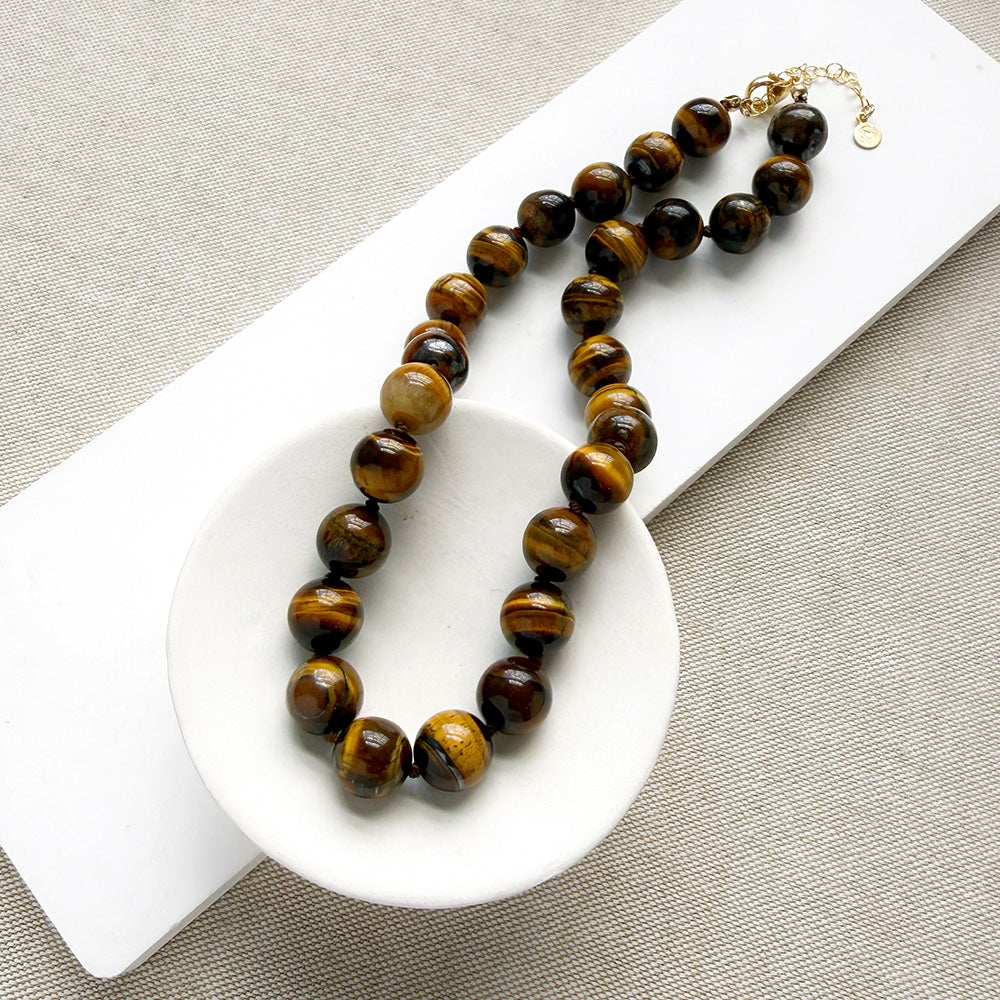 A beaded necklace made of polished tiger's eye beads in brown and gold tones, draped over a small white dish on a white rectangular board over a textured beige fabric background.