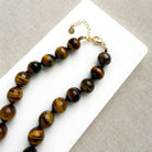 A beaded necklace made of polished tiger's eye beads in brown and gold tones, draped over a small white dish on a white rectangular board over a textured beige fabric background.