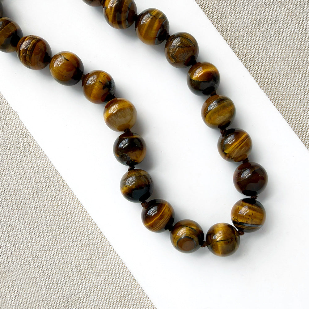 A beaded necklace made of polished tiger's eye beads in brown and gold tones, draped over a small white dish on a white rectangular board over a textured beige fabric background.