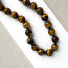 A beaded necklace made of polished tiger's eye beads in brown and gold tones, draped over a small white dish on a white rectangular board over a textured beige fabric background.