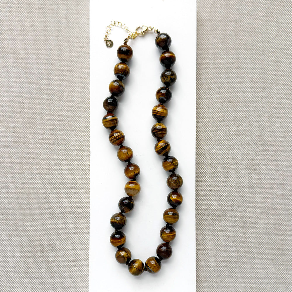 A beaded necklace made of polished tiger's eye beads in brown and gold tones, draped over a small white dish on a white rectangular board over a textured beige fabric background.