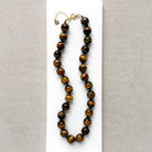 A beaded necklace made of polished tiger's eye beads in brown and gold tones, draped over a small white dish on a white rectangular board over a textured beige fabric background.