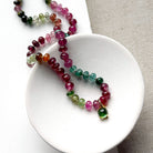 Smooth Watermelon Tourmaline beads in shades of pink, red, green, blue, and orange, featuring a small green tourmaline charm with gold detailing, displayed on a white ceramic dish.