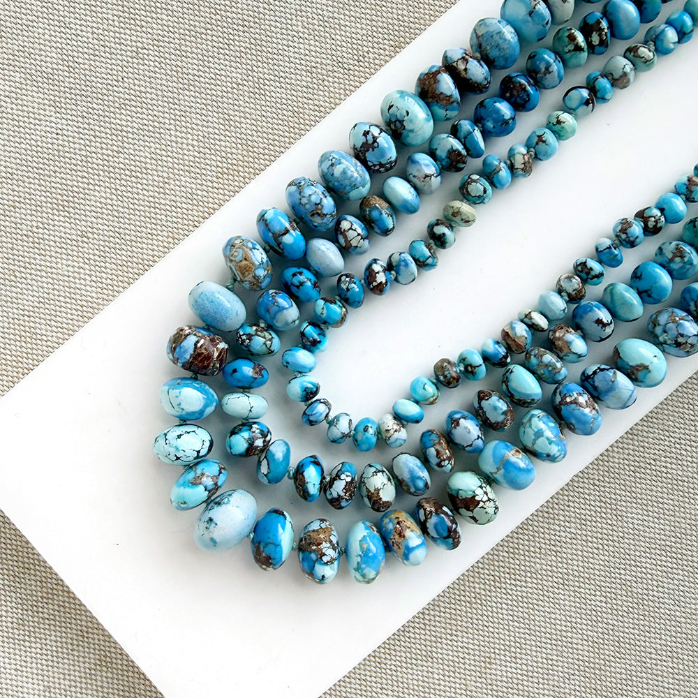 Three strands of polished turquoise beads in various sizes, showcasing shades of blue, light aqua, and brown matrix, arranged on a white rectangular card over a textured beige fabric background.