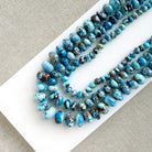 Three strands of polished turquoise beads in various sizes, showcasing shades of blue, light aqua, and brown matrix, arranged on a white rectangular card over a textured beige fabric background.