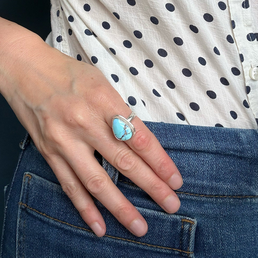 Hand wearing a Golden Hills turquoise ring with a polka dot shirt and blue jeans in the background