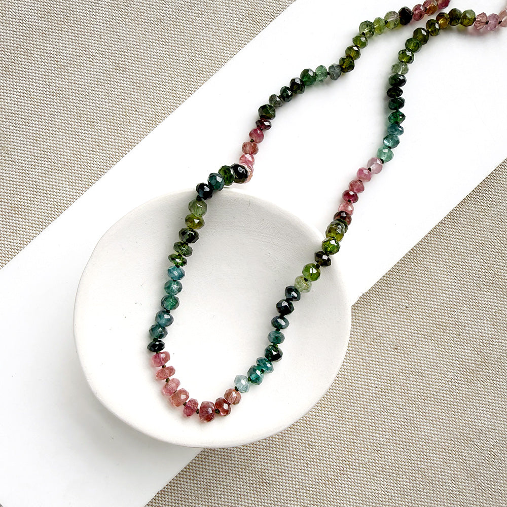 A beaded necklace made of Watermelon Tourmaline faceted round beads in shades of green, blue, and pink, draped over a small white dish on a textured beige fabric background.