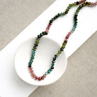 A beaded necklace made of Watermelon Tourmaline faceted round beads in shades of green, blue, and pink, draped over a small white dish on a textured beige fabric background.