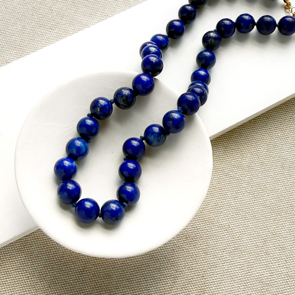 A beaded necklace made of polished dark blue lapis lazuli round beads, draped over a white ceramic dish on a textured beige fabric background.