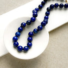 A beaded necklace made of polished dark blue lapis lazuli round beads, draped over a white ceramic dish on a textured beige fabric background.