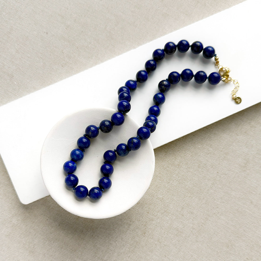 A beaded necklace made of dark blue lapis lazuli stones with a gold clasp, displayed on a small white dish over a white rectangular card on a beige fabric background.