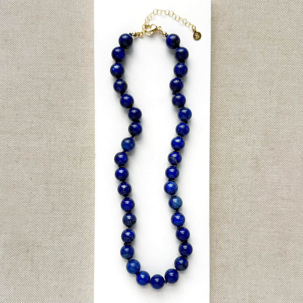 A beaded necklace made of polished dark blue lapis lazuli stones, displayed on a white card against a neutral beige textured fabric background. The necklace features a gold-tone clasp and extender chain.
