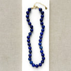 A beaded necklace made of polished dark blue lapis lazuli stones, displayed on a white card against a neutral beige textured fabric background. The necklace features a gold-tone clasp and extender chain.