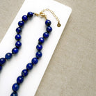 A beaded necklace made of polished dark blue lapis lazuli stones with a gold clasp and extender chain, displayed on a white rectangular card over a beige woven fabric background.