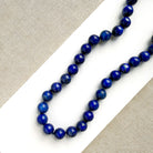 A strand of polished dark blue lapis lazuli ball beads arranged diagonally on a white surface with a beige textured fabric background.