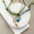 A layered gold-tone necklace set displayed on a white circular stand over a beige fabric background. The necklace features green and blue-green beaded strands with a teardrop-shaped blue-green stone pendant and a small gold fish charm.