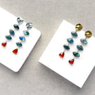 One  pair of gold and one pair of silver dangle earrings with marquis cut labradorite gems and a triangular hessian garnet, displayed on a white square card over a textured beige fabric background.