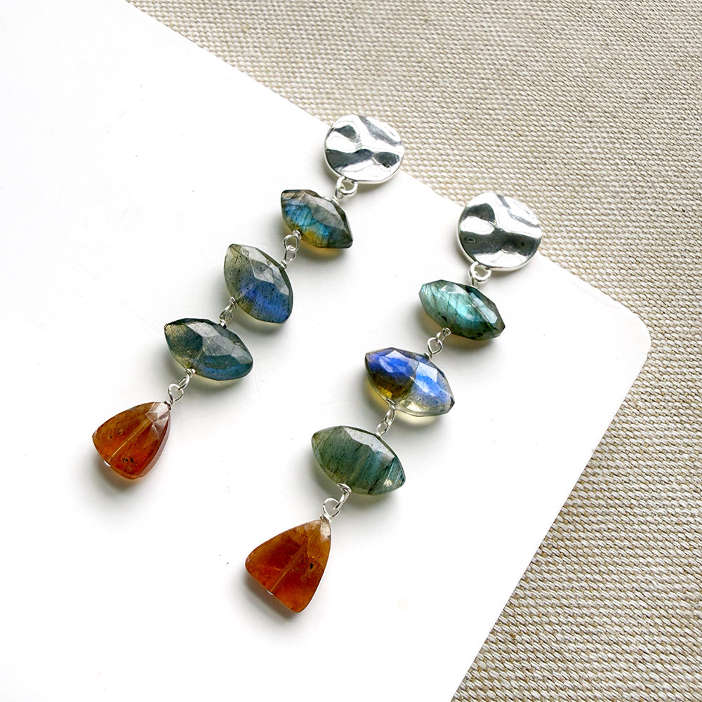 A pair of silver dangle earrings with marquis cut labradorite gems and a triangular hessian garnet, displayed on a white square card over a textured beige fabric background.