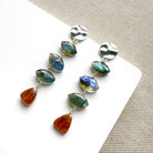 A pair of silver dangle earrings with marquis cut labradorite gems and a triangular hessian garnet, displayed on a white square card over a textured beige fabric background.