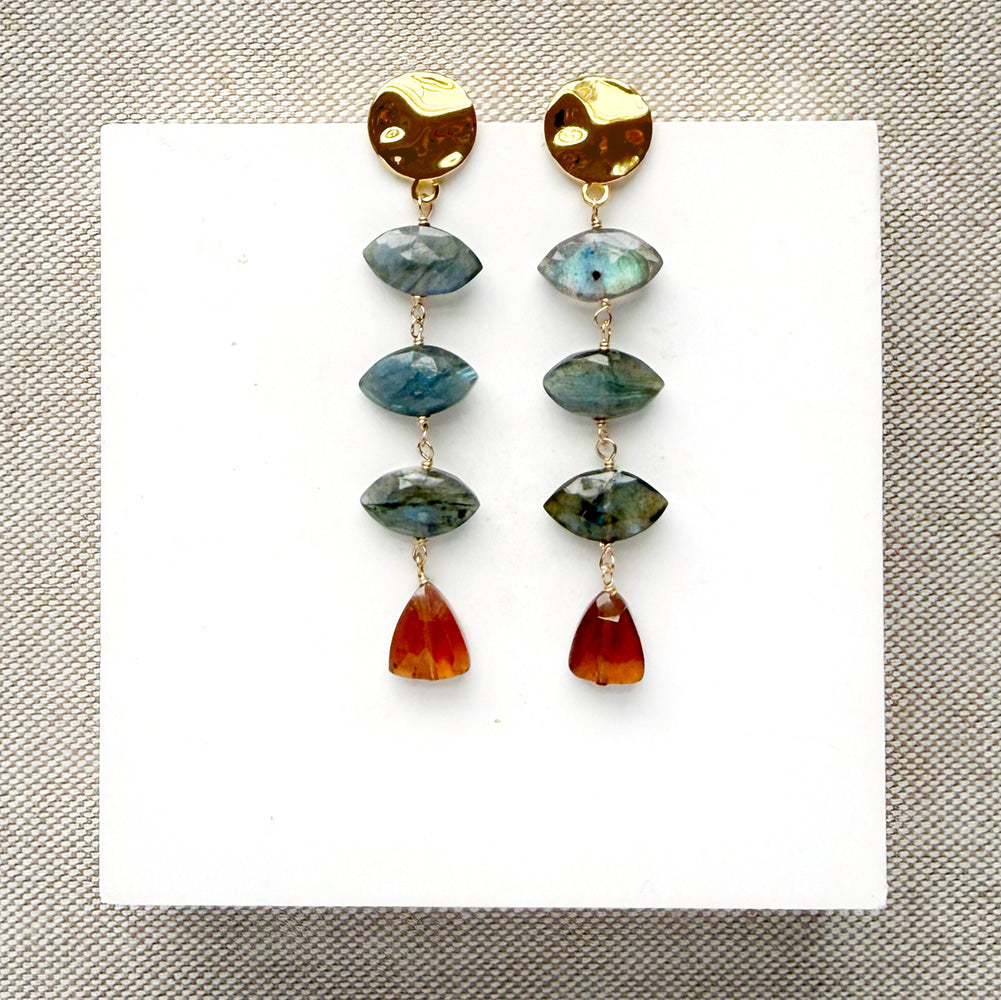 A pair of gold-tone dangle earrings with marquis cut labradorite gems and a triangular hessian garnet, displayed on a white square card over a textured beige fabric background.