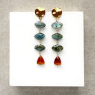 A pair of gold-tone dangle earrings with marquis cut labradorite gems and a triangular hessian garnet, displayed on a white square card over a textured beige fabric background.