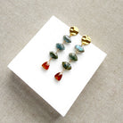 A pair of gold dangle earrings with marquis cut labradorite gems and a triangular hessian garnet, displayed on a white square card over a textured beige fabric background.