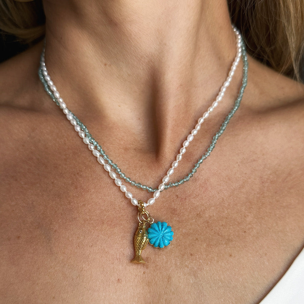 A close-up of a model wearing layered necklaces with white pearls and a light blue-green gemstones. The necklace features a gold-tone fish charm and a small howlite flower pendant.