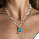 A close-up of a model wearing layered necklaces with white pearls and a light blue-green gemstones. The necklace features a gold-tone fish charm and a small howlite flower pendant.