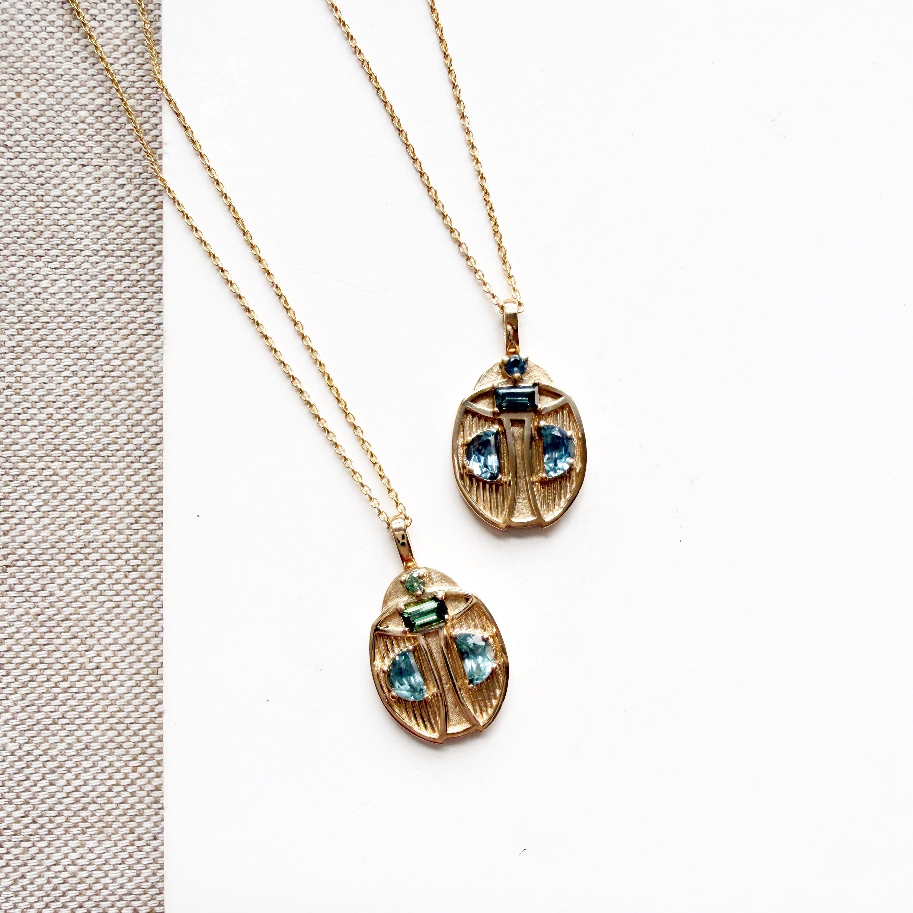 Two gold beetle-shaped pendant necklaces with blue gemstone accents, displayed on a white card over a textured beige fabric background.