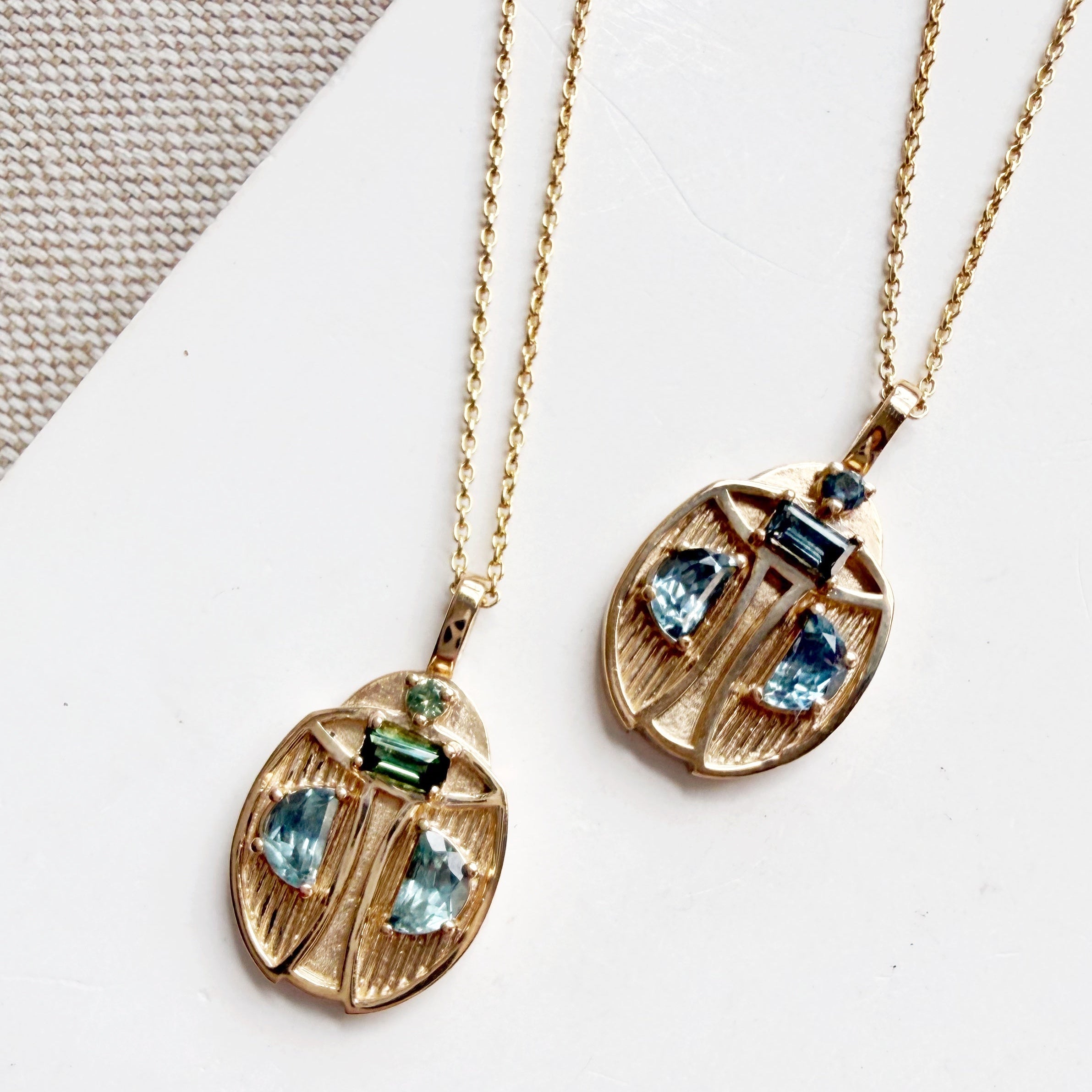 Two gold beetle-shaped pendant necklaces with blue and green gemstones, displayed on a white card over a textured beige fabric background.