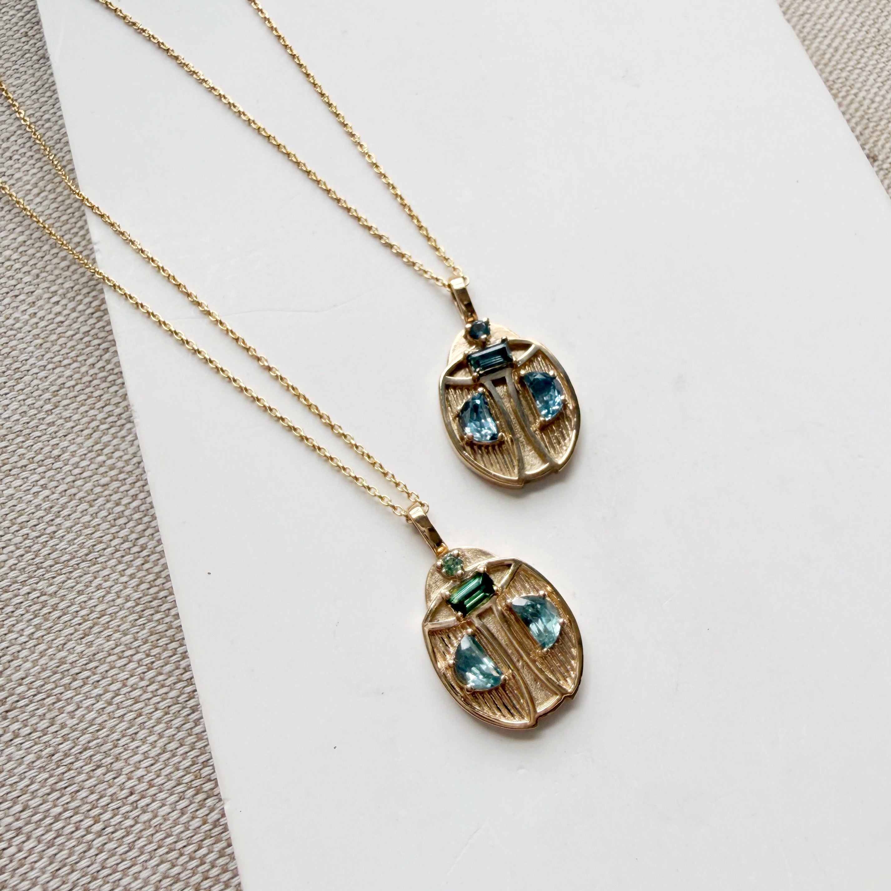 Two gold beetle-shaped pendant necklaces with blue gemstones, displayed on a white card over a textured beige fabric background.