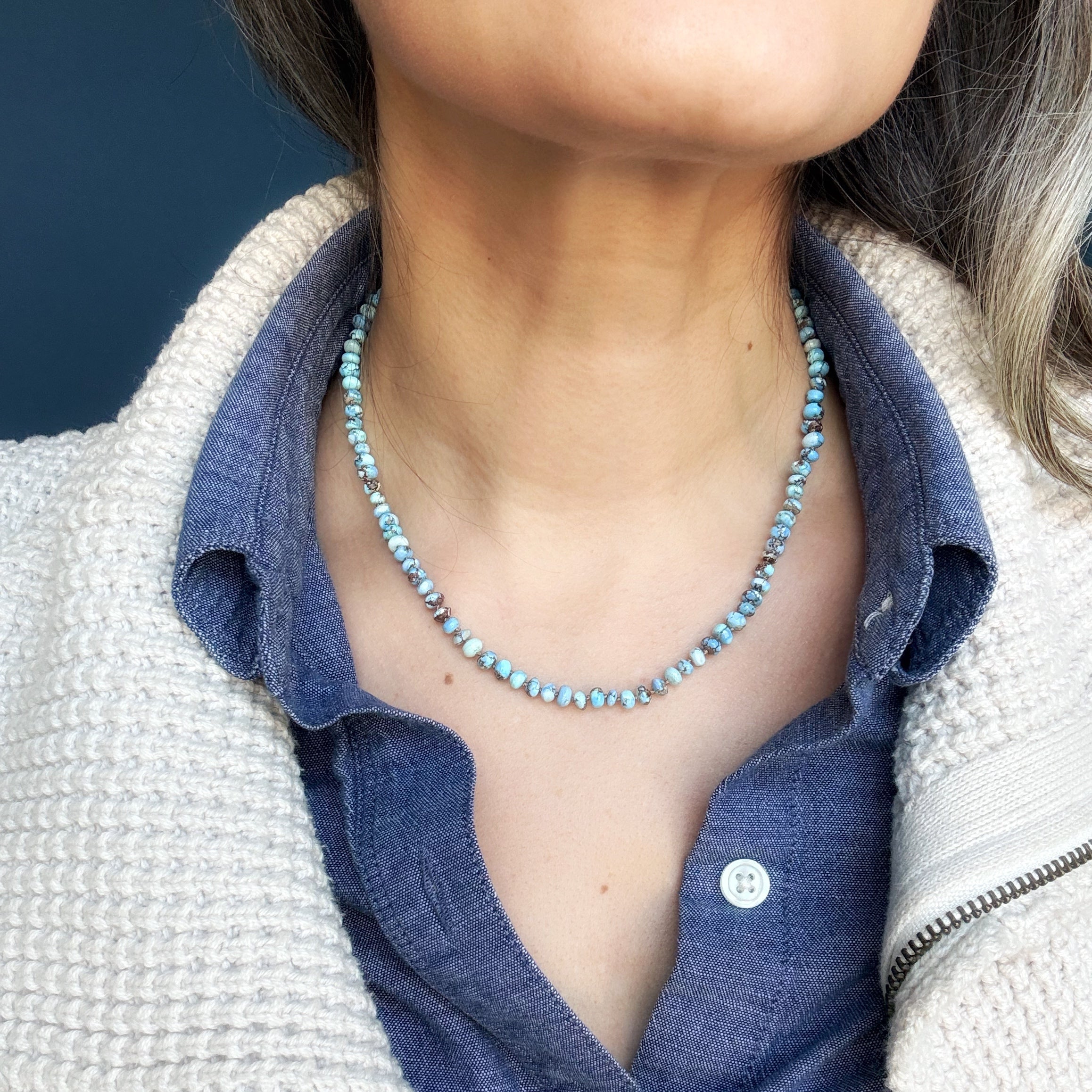 Person wearing a Golden Hills Turquoise beaded necklace with a blue shirt and beige jacket against a dark background