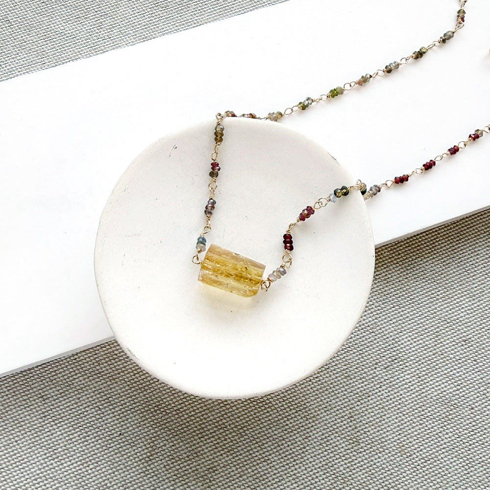 A multi-color sapphire necklace with a central imperial topaz pendant, displayed on a small white ceramic dish over a gray textured fabric background.