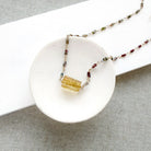 A multi-color sapphire necklace with a central imperial topaz pendant, displayed on a small white ceramic dish over a gray textured fabric background.