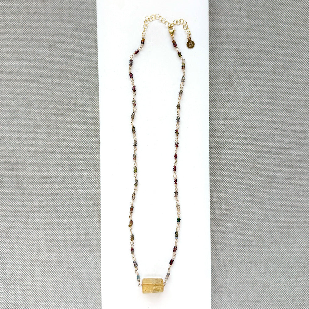 A multi-color sapphire necklace with a central imperial topaz pendant, displayed on a white background over a gray textured fabric background.