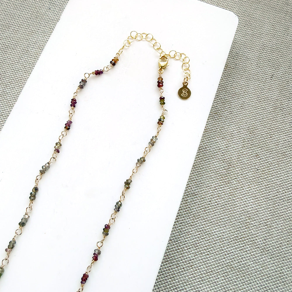 A multi-color sapphire necklace with a central imperial topaz pendant, displayed on a white background over a gray textured fabric background.
