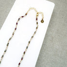 A multi-color sapphire necklace with a central imperial topaz pendant, displayed on a white background over a gray textured fabric background.