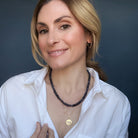 Woman wearing two necklaces, one with iolite gemstones and the other with a gold coin pendant, on a white shirt.