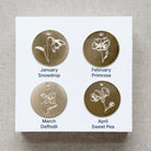 Four gold floral charms on a white card with month and flower name labels, on a textured beige background.
