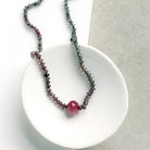 A necklace with small multicolored sapphire beads and a prominent oval pink sapphire at the center, displayed on a white ceramic dish against a light textured background.