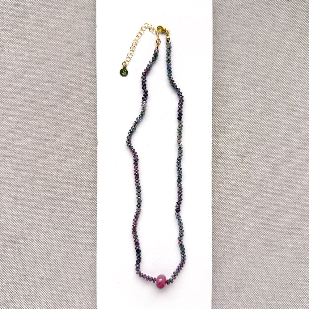 A necklace with small multicolored sapphire beads and a prominent oval pink sapphire at the center, displayed on a white ceramic dish against a light textured background.