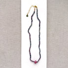 A necklace with small multicolored sapphire beads and a prominent oval pink sapphire at the center, displayed on a white ceramic dish against a light textured background.