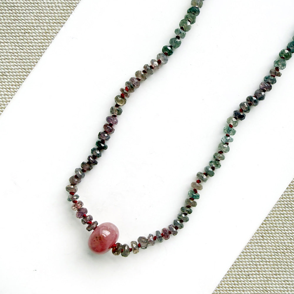 A necklace with small multicolored sapphire beads and a prominent oval pink sapphire at the center, displayed on a white dish against a light textured background.