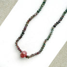 A necklace with small multicolored sapphire beads and a prominent oval pink sapphire at the center, displayed on a white dish against a light textured background.
