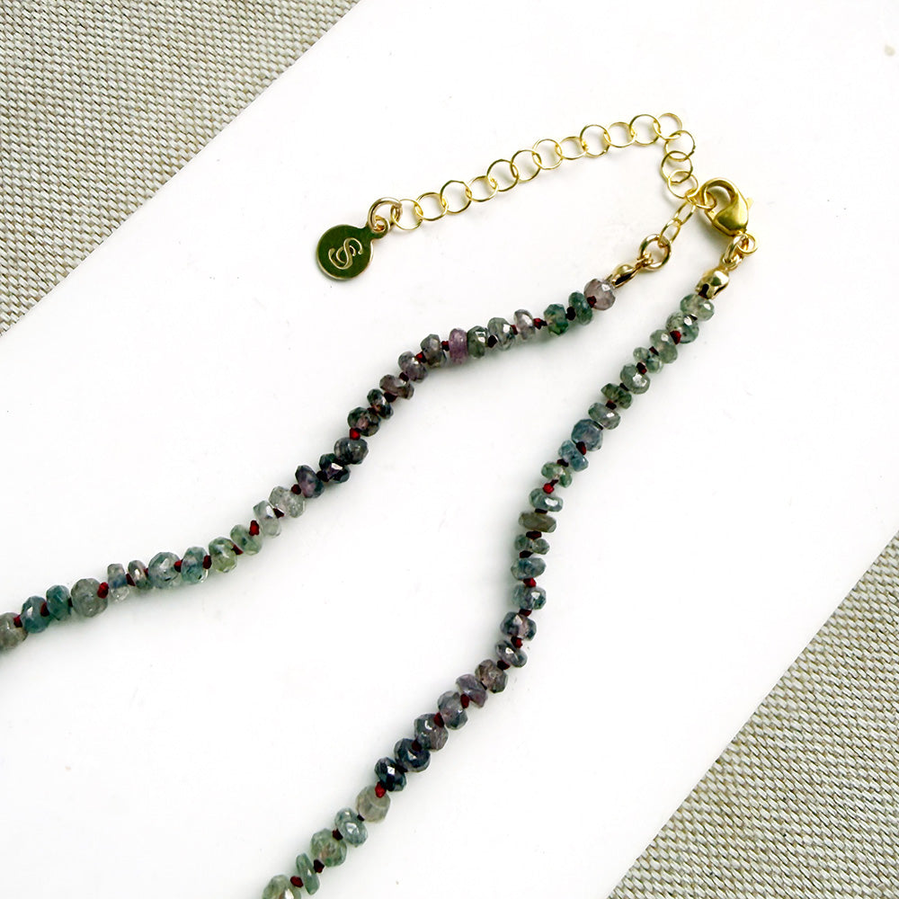A necklace with small multicolored sapphire beads and a prominent oval pink sapphire at the center, displayed on a white ceramic dish against a light textured background.