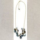 A gold chain necklace with multiple faceted labradorite gemstone charms in shades of blue, green, gray, and black, arranged on a white card over a beige woven fabric background.