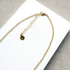 Close-up of a gold chain necklace with a lobster clasp, extension chain, and a small round engraved disc charm, laid on a white rectangular surface over a textured gray fabric background.