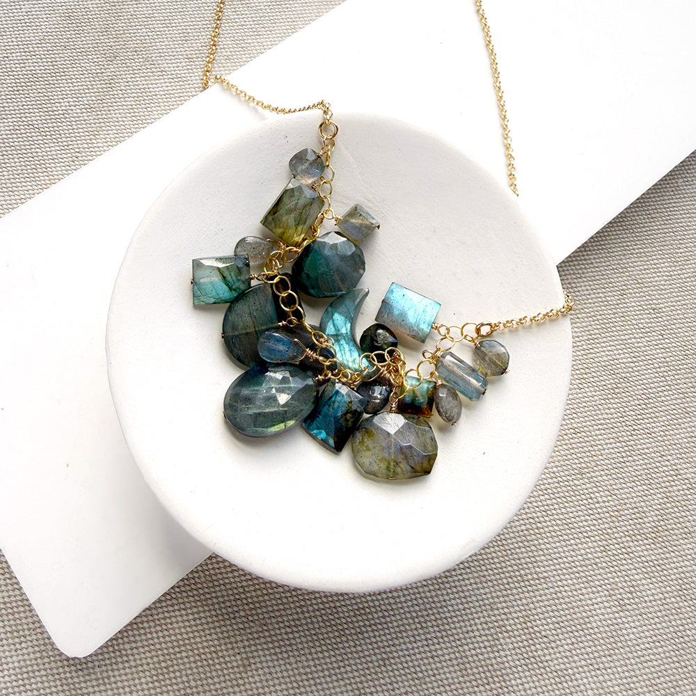 A gold chain necklace with a cluster of faceted labradorite gemstones in shades of blue, teal, and gray, displayed on a white ceramic dish over a textured beige fabric background.