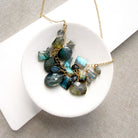 A gold chain necklace with a cluster of faceted labradorite gemstones in shades of blue, teal, and gray, displayed on a white ceramic dish over a textured beige fabric background.