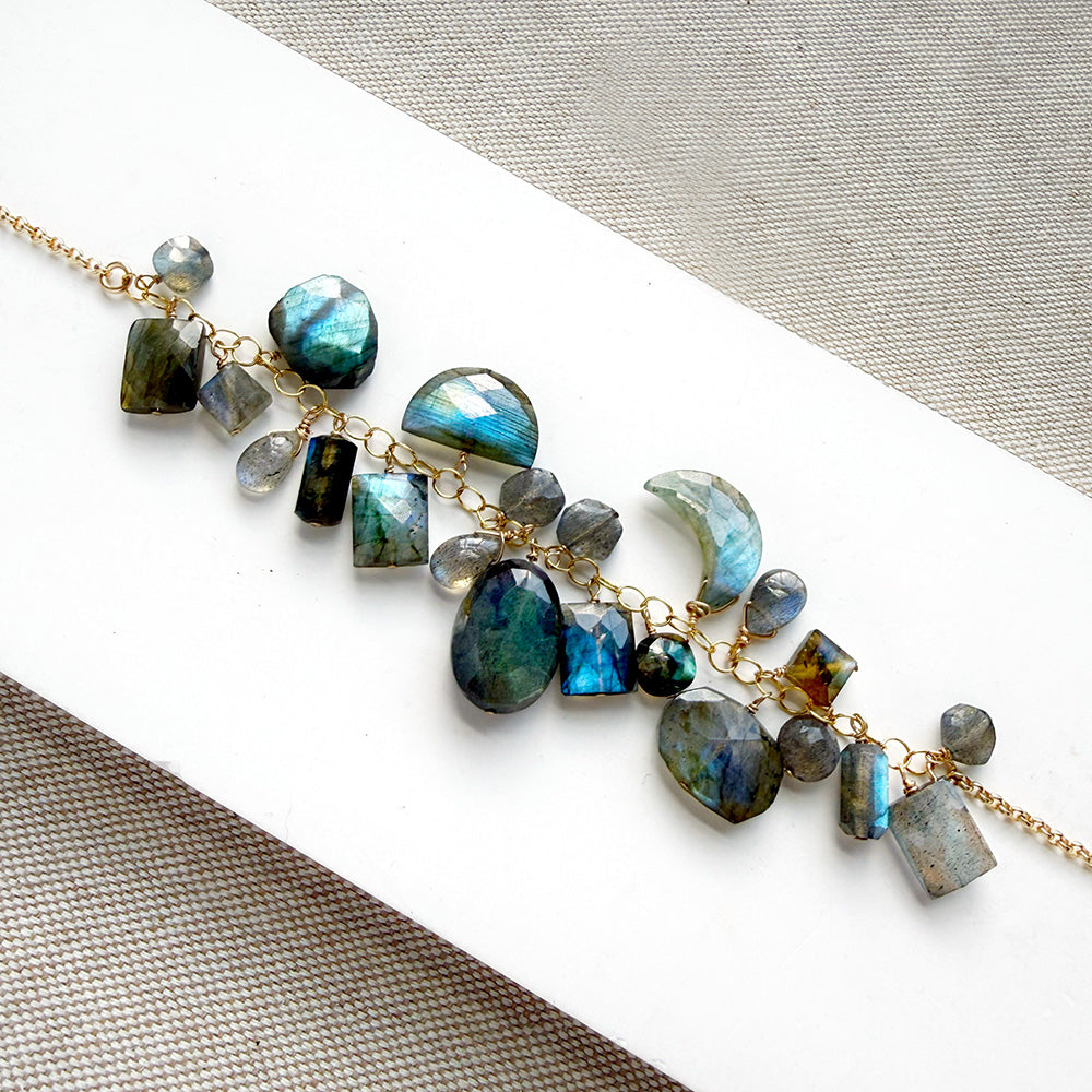 A gold chain necklace featuring natural labradorite gemstones in various shapes and sizes, displayed on a white rectangular card over a textured gray fabric background.
